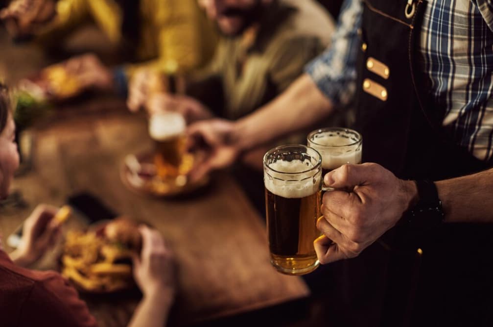 Close-up of hands serving beer mugs over a blurred food table