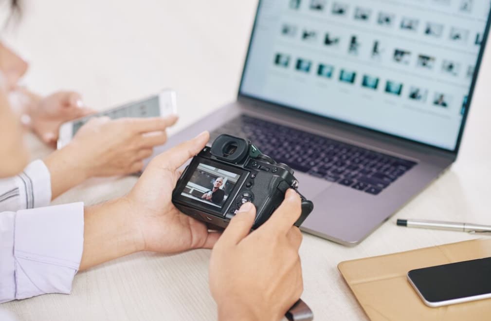 A person holding a camera reviews a photo of a man on its screen with a laptop nearby